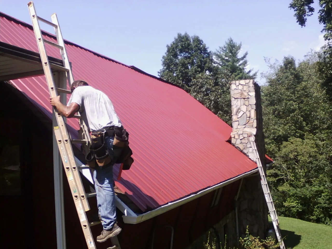 Roofer on a ladder beside a red metal roof and stone chimney — Soffit & Fascia Repair by Roofing Brothers in Wendell